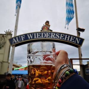 Una mano con braccialetti regge un grande boccale di birra di vetro di fronte a un cartello con la scritta 'AUF WIEDERSEHEN' durante un festival.