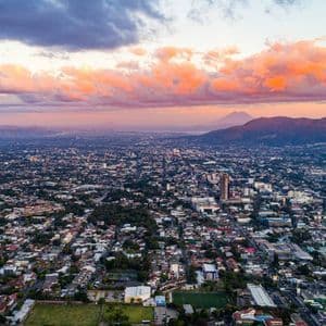 Eine Luftaufnahme einer weitläufigen Stadtlandschaft bei Sonnenuntergang, mit orangefarbenen Wolken darüber und Bergen am Horizont.