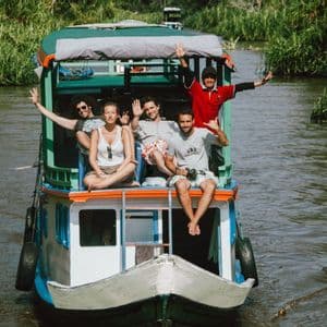 Un grupo de WeRoad sonríe y saluda desde la cubierta superior de un barco en un río rodeado de vegetación.