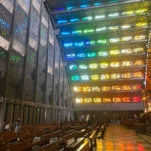 Das Innere einer modernen Betonkirche, in der farbiges Licht von Buntglasfenstern die Holzbänke beleuchtet.