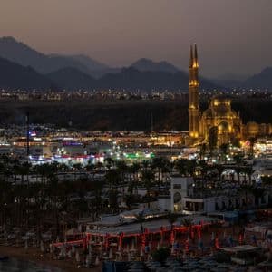 Una vista panoramica di una città costiera al tramonto, con una moschea illuminata e montagne sullo sfondo.