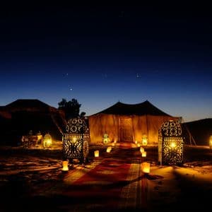 Lanterne illuminate e candele fiancheggiano un sentiero di tappeto rosso verso una tenda nel deserto sotto un cielo notturno stellato.