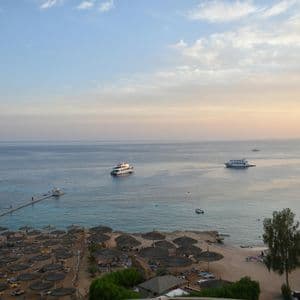 Una vista dall'alto di una spiaggia sabbiosa con ombrelloni di paglia e sedie a sdraio, con diversi yacht sul mare calmo al tramonto.