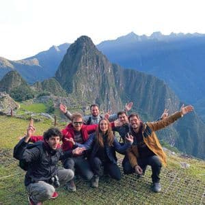 A WeRoad group trip posing for a photo on a grassy viewpoint with ancient ruins and steep green mountains in the background.
