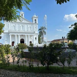 Eine weiße Kolonialkirche mit Säulen und Glockentürmen steht hinter einem Brunnen auf einem sonnigen Stadtplatz, von einem Park aus gesehen.