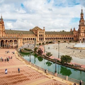 Una vista elevada de una gran plaza con un edificio semicircular ornamentado, un canal con puentes, una fuente central y gente caminando alrededor.