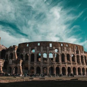 Un grand amphithéâtre antique en pierre se dresse sous un ciel sarcelle parsemé de nuages légers, avec une foule de gens rassemblée devant.
