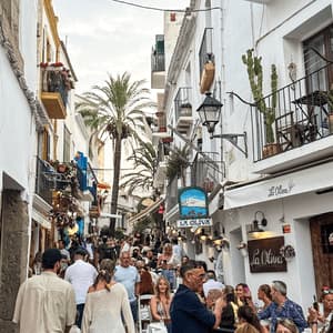 Una strada stretta e vivace, piena di persone che passeggiano e cenano nei ristoranti all'aperto, incastonata tra edifici bianchi.