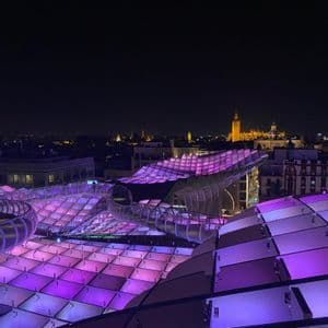 Una moderna estructura arquitectónica ondulada, iluminada con luces moradas por la noche, con vistas a un horizonte urbano que incluye la torre resplandeciente de una catedral.