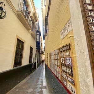 Un callejón estrecho con paredes amarillas, un pavimento de damero y azulejos cerámicos decorativos montados en un lado bajo un cielo azul.