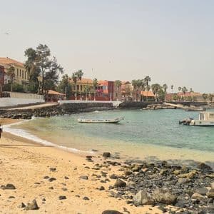 Ein sandiger, felsiger Strand an einer ruhigen Bucht mit vertäuten Booten, bunten Gebäuden und Palmen entlang der Uferpromenade.