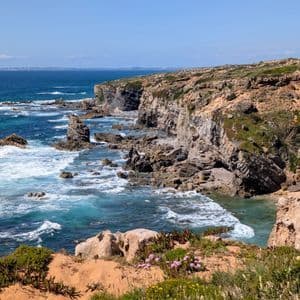 Eine zerklüftete Küste mit steilen Klippen und Brandungspfeilern, die unter einem klaren blauen Himmel von türkisfarbenen Wellen umspült wird.