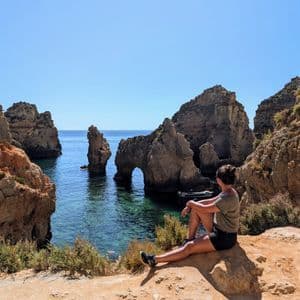 Eine Frau sitzt auf einer sonnigen Klippe und blickt auf ein ruhiges Meer mit großen Felsformationen und einem natürlichen Bogen.