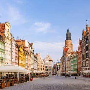 A wide cobblestone town square lined with colorful, historic buildings and outdoor cafes under a clear blue sky, with a tall church tower in the background.
