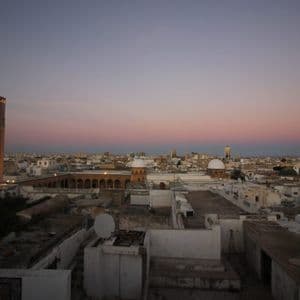 Un minaret illuminé domine un paysage urbain de bâtiments blancs et de toits plats sous un ciel rose et violet au crépuscule.