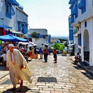 Un homme en tenue traditionnelle marche dans une rue pavée animée bordée d'étals de marché et de bâtiments blanchis à la chaux avec des garnitures bleues.