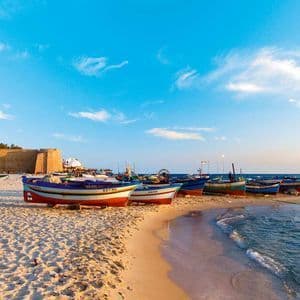 Des bateaux de pêche colorés sont alignés sur une plage de sable près de la mer, avec une grande forteresse en pierre en arrière-plan.