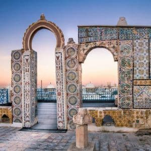 Une terrasse sur le toit avec des arches ornées et des mosaïques colorées, surplombant un paysage urbain avec un minaret au coucher du soleil.