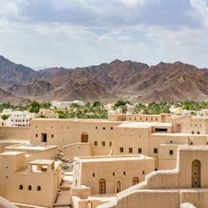 Ein historisches Dorf mit sandfarbenen Gebäuden und einem runden Turm, mit einer grünen Oase und schroffen Bergen im Hintergrund.