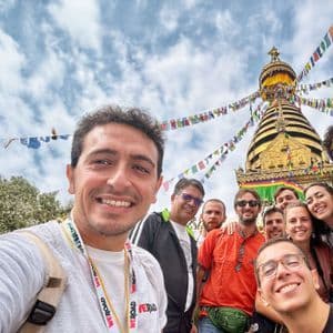 Un gruppo WeRoad si scatta un selfie sorridente davanti a una stupa dorata, con bandiere di preghiera colorate che sventolano sotto un cielo nuvoloso.