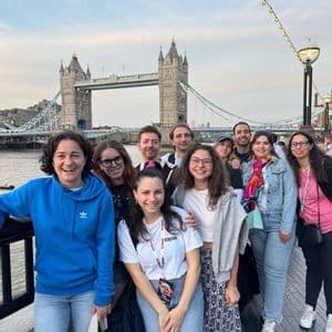 Un gruppo WeRoad in viaggio posa sorridendo per una foto su un lungomare, con un grande ponte storico visibile oltre il fiume.