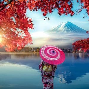 Una persona in kimono con un ombrellino rosa in riva a un lago, osserva una montagna innevata incorniciata da foglie autunnali rosse.