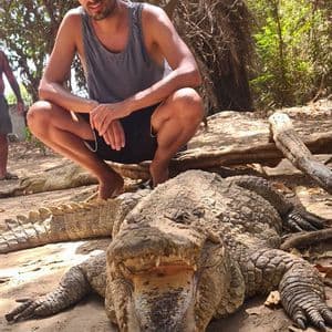 Ein Mann hockt auf einer WeRoad-Gruppenreise barfuß hinter einem großen Krokodil mit geöffnetem Maul auf sandigem Boden.