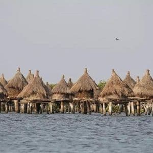 Ein Pfahlbautendorf mit konischen strohgedeckten Hütten steht in ruhigem Wasser unter einem dunstigen Himmel.