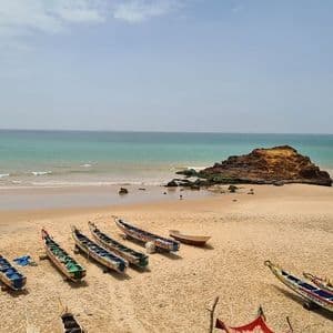 Mehrere bunte Pirogenboote liegen an einem breiten Sandstrand neben dem türkisfarbenen Meer, mit einer felsigen Insel direkt vor der Küste.