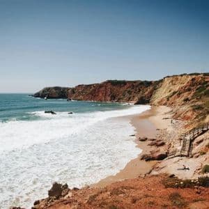 Meereswellen mit weißem Schaum branden an einem Sandstrand, der sich zwischen rotbraunen Klippen unter einem klaren blauen Himmel schmiegt.