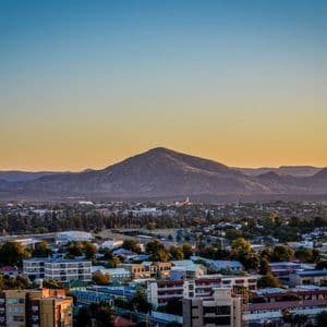 Eine Luftaufnahme einer Stadt mit Gebäuden und Bäumen, eingebettet am Fuße von Bergen unter einem goldenen Sonnenuntergangshimmel.