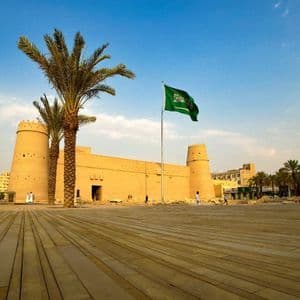 La bandiera dell'Arabia Saudita sventola davanti a una grande fortezza color sabbia con palme e un'ampia piazza.