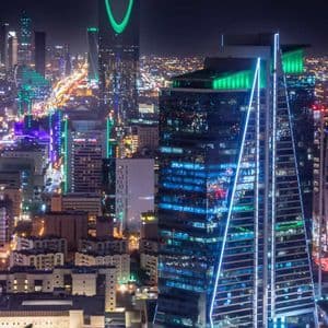Un vaste panorama urbain nocturne, avec des gratte-ciel illuminés en vert, bleu et rouge, et les traînées lumineuses des véhicules dans les rues en contrebas.