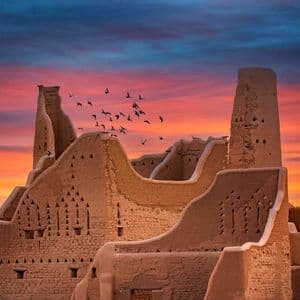 Un vol d'oiseaux survole d'anciennes ruines en briques de terre avec des tours, sous un ciel de coucher de soleil coloré.