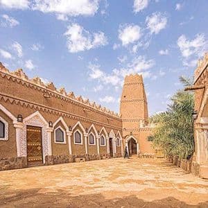 Un bâtiment traditionnel en pisé avec une tour et des arches blanches décoratives se dresse dans une cour en pierre sous un ciel bleu.