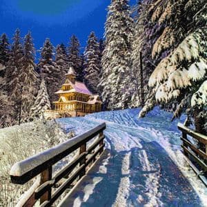 Una capilla de madera, iluminada por la noche, se asienta en un bosque nevado de pinos, con un puente cubierto de nieve en primer plano.