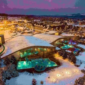Una vista aérea de un gran complejo de spa con piscinas turquesa iluminadas, situado en un pueblo cubierto de nieve al atardecer.