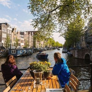 Deux personnes assises à une table en bois sur une terrasse, regardant un canal de la ville avec des bateaux et des bâtiments.