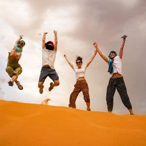 Un voyage de groupe WeRoad de quatre personnes sautant en l'air sur une dune de sable orange sous un ciel partiellement nuageux.