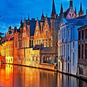 Des bâtiments illuminés aux flèches gothiques bordent un canal au crépuscule, leurs reflets chatoyant sur l'eau.