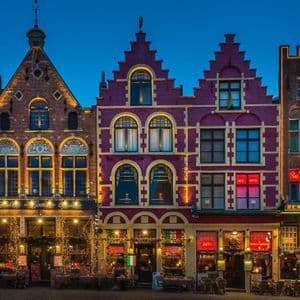 Une rangée de bâtiments historiques colorés avec des pignons à redents, illuminés par des lumières de Noël festives au crépuscule.