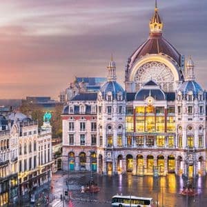Un bâtiment orné et en forme de dôme, brillamment éclairé au crépuscule sur une place de ville, avec des rues mouillées reflétant les lumières de l'architecture environnante.