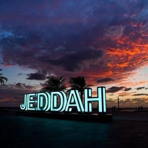 Une enseigne lumineuse épelant 'JEDDAH' sur un front de mer avec des palmiers sous un ciel de coucher de soleil coloré.