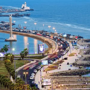 Vue aérienne d'une ville côtière animée au crépuscule, avec de la circulation sur une route courbée le long d'une lagune et un grand monument.