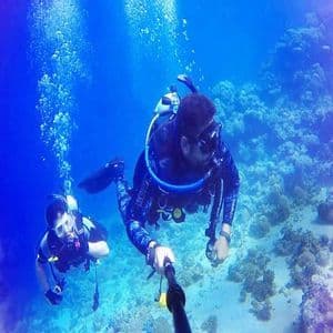 Deux personnes d'un voyage de groupe WeRoad faisant de la plongée sous-marine, l'une prenant une photo avec une perche à selfie au-dessus d'un récif corallien.
