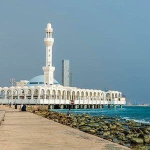 Une mosquée blanche avec un minaret et un dôme bleu se dresse sur une jetée au-dessus de la mer, avec un littoral rocheux et des bâtiments urbains modernes en arrière-plan.