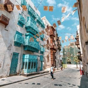 Un homme avec un sac à dos se tient dans une rue étroite, levant les yeux vers des bâtiments historiques avec des balcons en bois turquoises et marron.