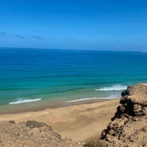 Una vista desde un acantilado rocoso con vistas a una playa de arena con gente nadando en el océano turquesa bajo un cielo azul claro.