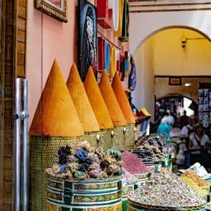 Tall cones of orange spices and colorful bins of dried herbs and flowers on display at an outdoor market stall.