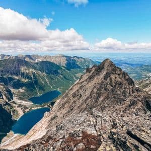 A panoramic view from a rocky mountain peak overlooking a vast mountain range with two blue alpine lakes in a valley below.